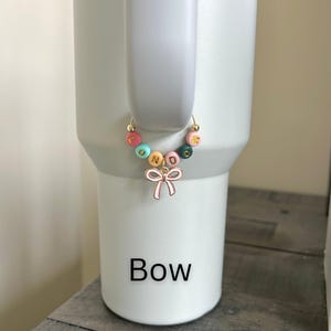 May include: A colorful beaded charm with the word "FOND" spelled out in gold letters, attached to a gold hoop. A pink bow charm hangs below the beads. The word "Bow" is printed in black below the charm.