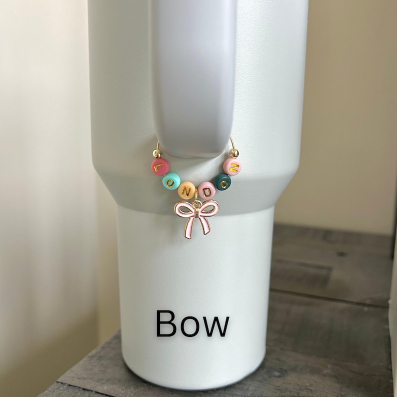 May include: A colorful beaded charm with the word "FOND" spelled out in gold letters, attached to a gold hoop. A pink bow charm hangs below the beads. The word "Bow" is printed in black below the charm.