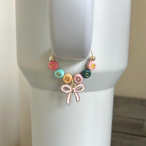 May include: A gold-colored charm bracelet with colorful beads spelling "LONDON" and a pink bow charm. The beads are in shades of pink, blue, yellow, and green. The bracelet is attached to a white tumbler.