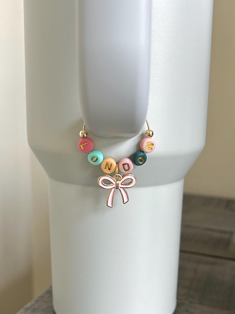 May include: A gold-colored charm bracelet with colorful beads spelling "LONDON" and a pink bow charm. The beads are in shades of pink, blue, yellow, and green. The bracelet is attached to a white tumbler.