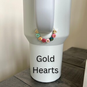 May include: A white tumbler with a gold heart-shaped charm and a beaded bracelet spelling "IANN" in colorful beads. The text "Gold Hearts" is printed in black on the tumbler.
