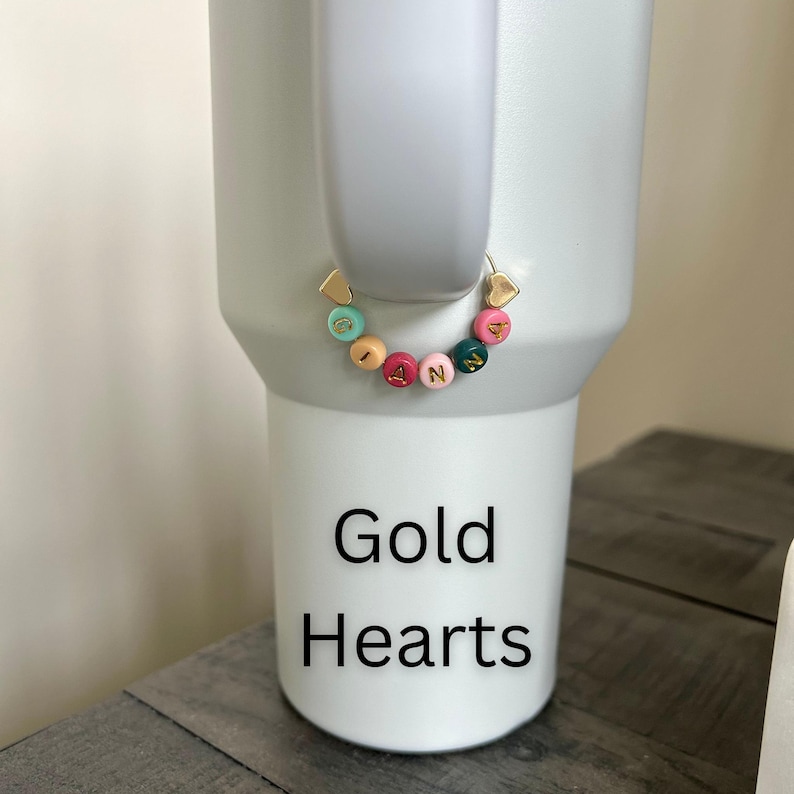 May include: A white tumbler with a gold heart-shaped charm and a beaded bracelet spelling "IANN" in colorful beads. The text "Gold Hearts" is printed in black on the tumbler.