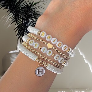 May include: A set of white and gold beaded bracelets with the word "BRIDE" and the numbers "091325" spelled out. A charm with the letter "H" hangs from the bottom of the bracelets.