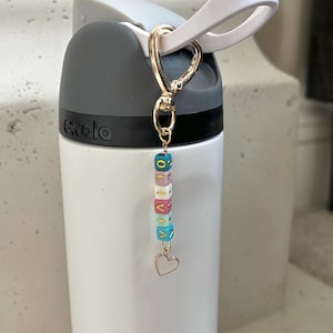 May include: A white water bottle with a grey top and a gold-coloured clip holding a charm. The charm has colourful lettered beads spelling "OLIVIA" and a small heart pendant. The bottle is on a white surface.