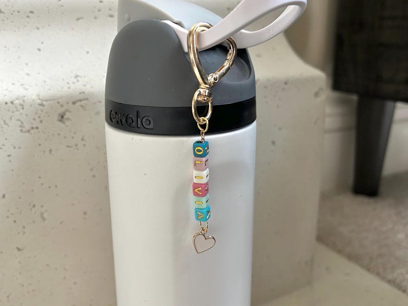 May include: A white water bottle with a grey top and a gold-coloured clip holding a charm. The charm has colourful lettered beads spelling "OLIVIA" and a small heart pendant. The bottle is on a white surface.