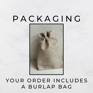 May include: A beige burlap bag with a drawstring closure. The bag is sitting on a white marble surface. The text "PACKAGING" is above the bag and the text "YOUR ORDER INCLUDES A BURLAP BAG" is below the bag.