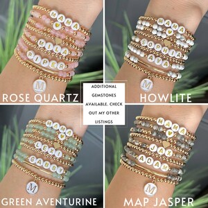 May include: A set of gold bead bracelets with different gemstone beads and personalized name charms. The bracelets are stacked on a wrist and include rose quartz, howlite, green aventurine, and map jasper beads. Each bracelet has a gold charm with a letter 'M' on it.