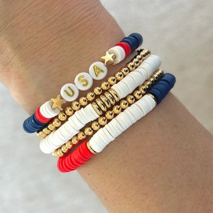 May include: A stack of bracelets in red, white, and blue with gold beads and a charm that says "USA" with a gold star.
