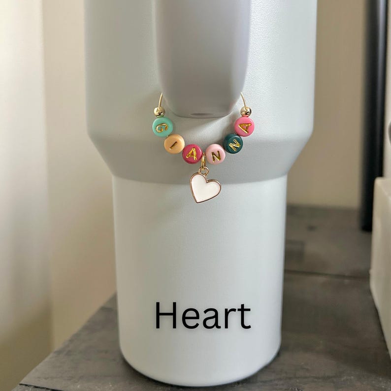 May include: A gold hoop earring with colorful letter beads spelling "GIANNA" and a white heart charm. The earring is displayed on a white tumbler. The word "Heart" is printed in black at the bottom of the tumbler.