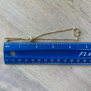 May include: A gold-colored bracelet with a delicate chain and a sliding bead for adjustment. The bracelet features small decorative elements, including a charm with a small black and white design. The bracelet is approximately 5 inches long.