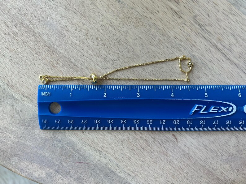 May include: A gold-colored bracelet with a delicate chain and a sliding bead for adjustment. The bracelet features small decorative elements, including a charm with a small black and white design. The bracelet is approximately 5 inches long.