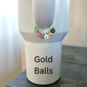 May include: A gold hoop earring with colorful letter beads spelling "LIVE". The earring is displayed on a light gray tumbler. The tumbler has the words "Gold Balls" printed on it in black.