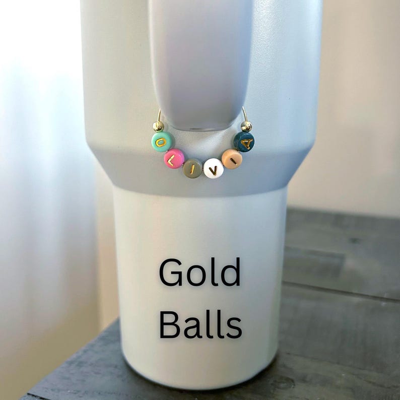 May include: A gold hoop earring with colorful letter beads spelling "LIVE". The earring is displayed on a light gray tumbler. The tumbler has the words "Gold Balls" printed on it in black.