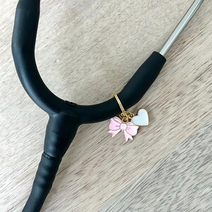 May include: A black stethoscope with a gold charm featuring a pink bow and a white heart. The stethoscope is on a light wood surface. Medical-themed accessory.