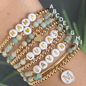 May include: A stack of gold bead bracelets with white letter beads spelling out the names "MAMA", "LUCAS", "GAVIN", and "CAS". The bracelets are on a person's wrist and there is a gold charm with the letter "M" hanging from the bottom of the stack.