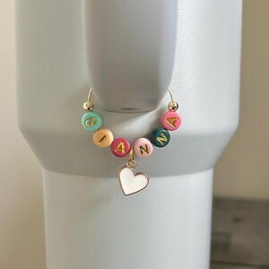 May include: A gold-toned charm with the name "IANNA" spelled out in colorful round beads. A white heart charm hangs below the name. The charm is attached to a gold-toned hoop, likely for a tumbler or cup.