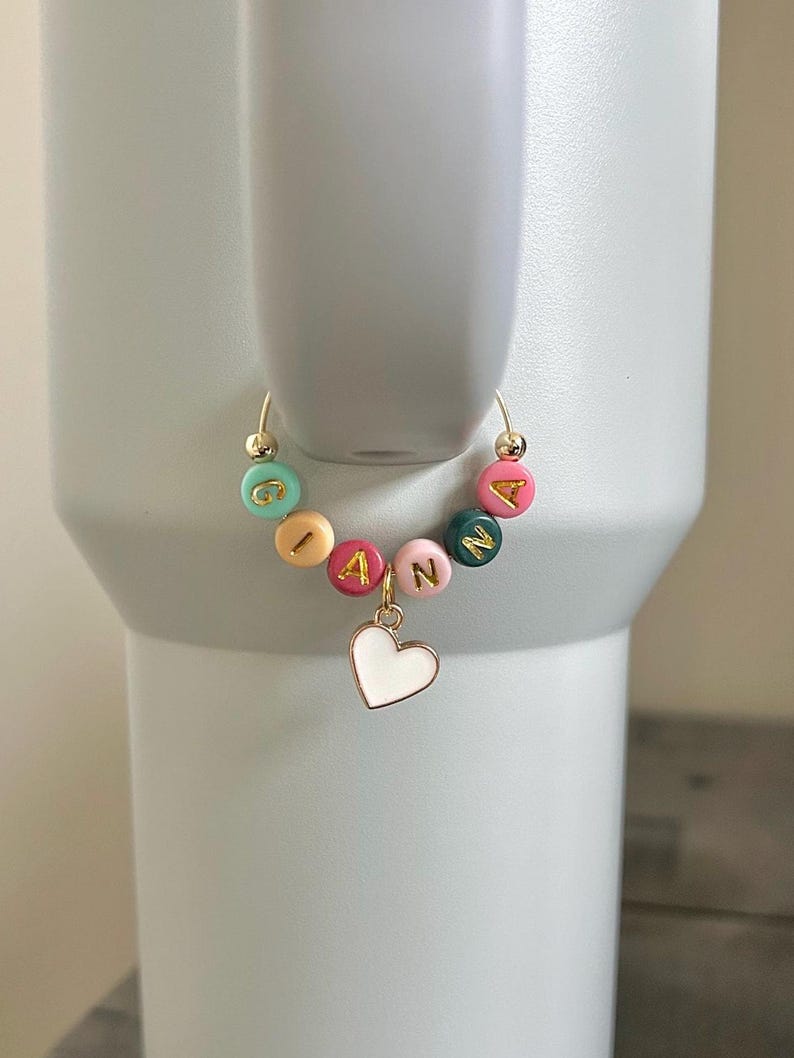 May include: A gold-toned charm with the name "IANNA" spelled out in colorful round beads. A white heart charm hangs below the name. The charm is attached to a gold-toned hoop, likely for a tumbler or cup.