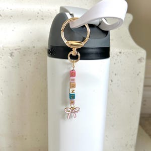May include: A white water bottle with a gold-coloured keychain. The keychain has colourful lettered blocks spelling "GIANNA" and a pink bow charm. The bottle has a black top and a white handle.
