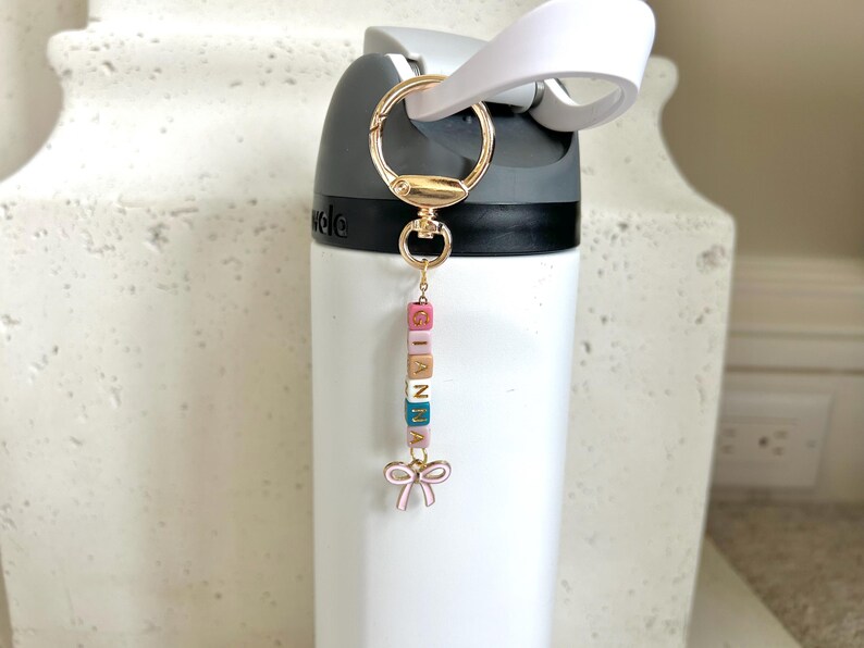 May include: A white water bottle with a gold-coloured keychain. The keychain has colourful lettered blocks spelling "GIANNA" and a pink bow charm. The bottle has a black top and a white handle.