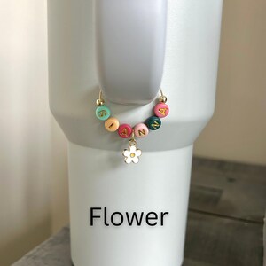 May include: A gold-colored tumbler charm with colorful letter beads spelling "IANN" and a white flower charm. The charm is attached to a white tumbler. The word "Flower" is printed in black at the bottom.