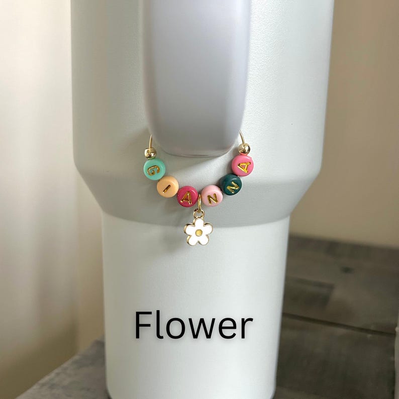 May include: A gold-colored tumbler charm with colorful letter beads spelling "IANN" and a white flower charm. The charm is attached to a white tumbler. The word "Flower" is printed in black at the bottom.