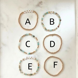 May include: Six gold bracelets with different charms. The charms include the words "MAMA", "BYB", "LUCAS", and a heart. The bracelets are made of gold beads and are strung with light blue and white stones.