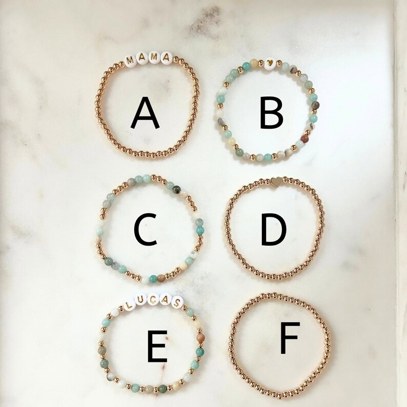 May include: Six gold bracelets with different charms. The charms include the words "MAMA", "BYB", "LUCAS", and a heart. The bracelets are made of gold beads and are strung with light blue and white stones.