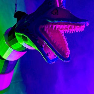 Pode incluir: Escultura colorida e estilizada de uma cabeça de dinossauro com a boca aberta, revelando dentes afiados. A escultura apresenta tons de azul, verde e rosa, com um fundo escuro. A imagem é iluminada por luzes de néon.