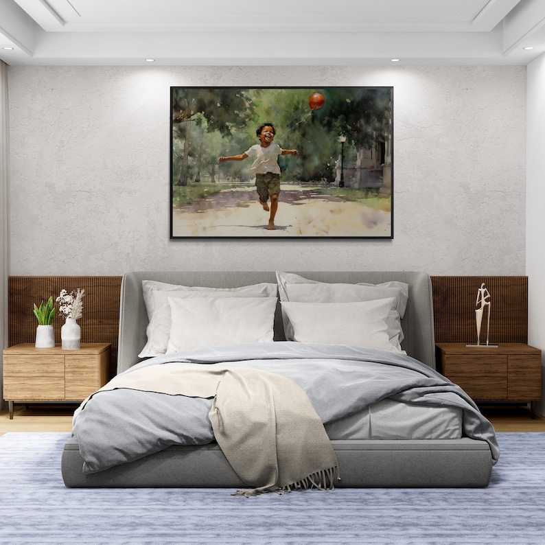 May include: A bedroom interior with a gray upholstered bed with white bedding and a throw blanket. A framed watercolor painting of a young boy running with a red ball in a park hangs on the wall above the bed. Two wooden nightstands with lamps are on either side of the bed.