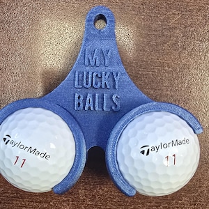 Könnte beinhalten: Ein blauer 3D-gedruckter Golfballhalter mit dem Text "MY LUCKY BALLS" und zwei weißen TaylorMade Golfbällen. Jeder Ball hat die Nummer 11 in Rot. Der Halter hat eine Schlaufe zum Aufhängen.