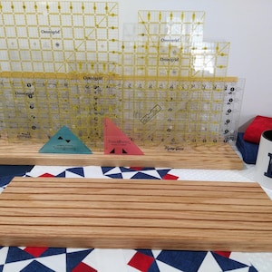 May include: A set of four clear plastic rulers with yellow markings and the word "Omnigrid" printed on them. The rulers are stacked on a wooden stand with a wooden cutting board in front of them. The cutting board has a striped pattern and is made of light wood.