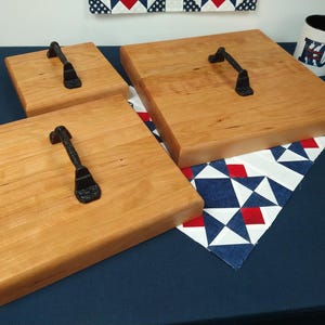 May include: Three wooden serving trays with black metal handles. The trays are different sizes and are arranged on a blue surface with a red, white, and blue patterned fabric underneath.