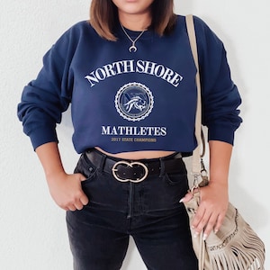 Può includere: Una felpa blu navy corta con il testo "NORTH SHORE" in bianco sopra uno stemma blu e oro con il testo "MATHLETES 2017 STATE CHAMPIONS". La felpa è indossata con jeans neri e una borsa beige con frange.