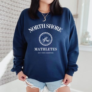 Può includere: Una felpa blu navy con testo bianco che recita "North Shore" in un arco sopra un logo circolare con un leone e il testo "Mathletes 2017 State Champions".