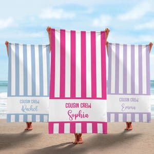 May include: Three beach towels with vertical stripes in blue, pink, and lavender, held up on a beach. Each towel has a white band with "COUSIN CREW" and a name printed on it. The towels are on a sandy beach with the ocean in the background.