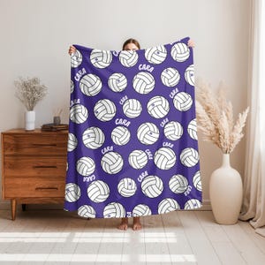 May include: A purple blanket featuring a repeating pattern of white volleyballs. Each volleyball has the name "CARA" and the number "10" printed on it. The blanket is held up by a person.