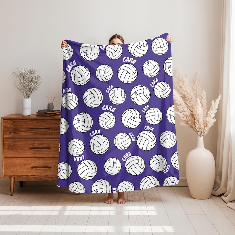 May include: A purple blanket featuring a repeating pattern of white volleyballs. Each volleyball has the name "CARA" and the number "10" printed on it. The blanket is held up by a person.