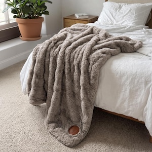 May include: A plush, taupe-colored faux fur throw blanket draped over a white bed. The blanket features a thick, shaggy texture and a circular, smooth brown leather-like patch detail near the bottom edge, adding a distinct contrast to the fabric.