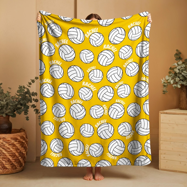 May include: Yellow blanket with a repeating pattern of white volleyballs and the name "RACHEL" in a white font. The blanket is held up by a person.