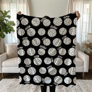 May include: Black blanket featuring a pattern of white volleyballs with the name "OLIVIA" printed on each. The volleyballs have a detailed design, and the blanket is held up in front of a neutral background.