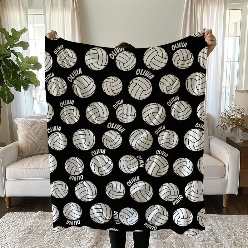 May include: Black blanket featuring a pattern of white volleyballs with the name "OLIVIA" printed on each. The volleyballs have a detailed design, and the blanket is held up in front of a neutral background.