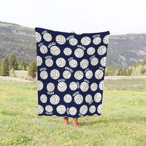 May include: Navy blue blanket with a repeating pattern of white volleyballs and the name "Rachel" in a white font. The blanket is held up in front of a person in a field with a mountain backdrop.