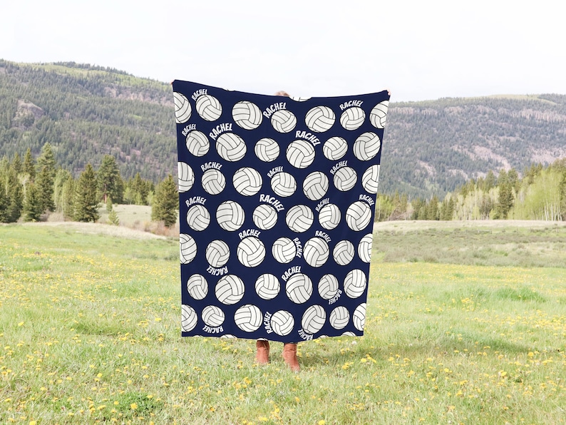 May include: Navy blue blanket with a repeating pattern of white volleyballs and the name "Rachel" in a white font. The blanket is held up in front of a person in a field with a mountain backdrop.
