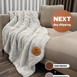 May include: A plush, elegant gray blanket draped over a beige sofa. The blanket has a textured, faux fur appearance. A brown leather patch with the name "Mrs. Brown" and a date is sewn on the blanket. The color options are dark brown and elegant gray.