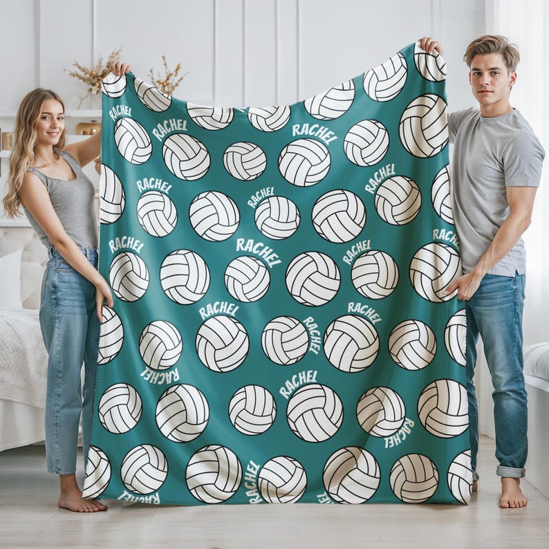 May include: A teal blanket featuring a pattern of white volleyballs and the name "RACHEL" in white. The blanket is held up by two people. The volleyballs are evenly spaced across the blanket.