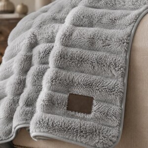 May include: A light gray faux fur throw blanket featuring a textured, horizontal ribbed pattern. The blanket has a smooth, finished edge and a rectangular brown faux leather patch sewn onto the lower corner of the fabric surface.