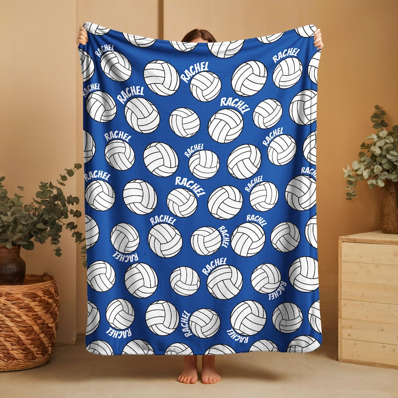 May include: A blue blanket featuring a pattern of white volleyballs. Each volleyball has the name "RACHEL" printed on it. The blanket is held up, showcasing the design and color.