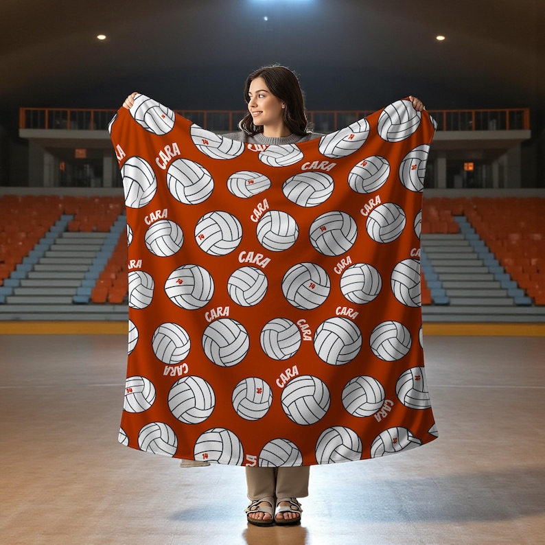 May include: A red blanket featuring a pattern of white volleyballs with the word "CARA" printed on each ball. The blanket is held up in front of a person in a sports arena.
