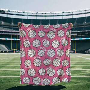 May include: Pink blanket featuring a repeating pattern of white volleyballs and the name "RACHEL" in a circular design. The blanket is held up against a backdrop of a green field and a stadium.