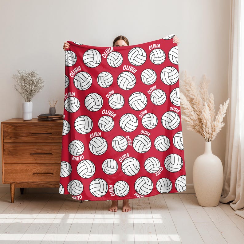 May include: Red blanket with a repeating pattern of white volleyballs and the name "Olivia" in white. The blanket is held up, showcasing the design. The blanket is a sports-themed item.